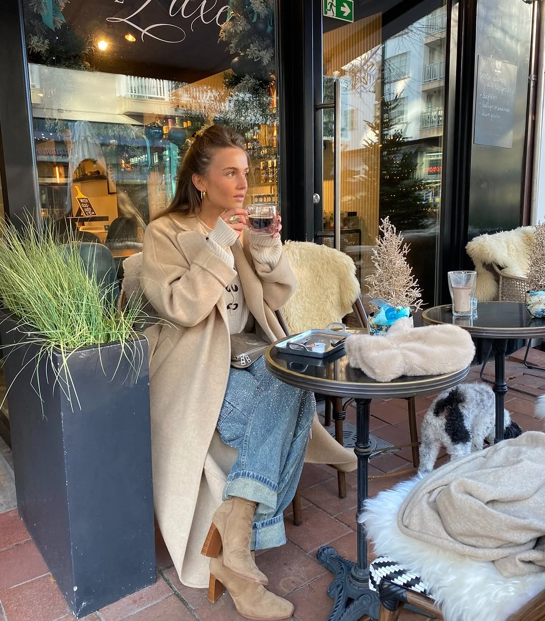 Long beige coat, cuffed blue jeans, tan boots, and gray bag