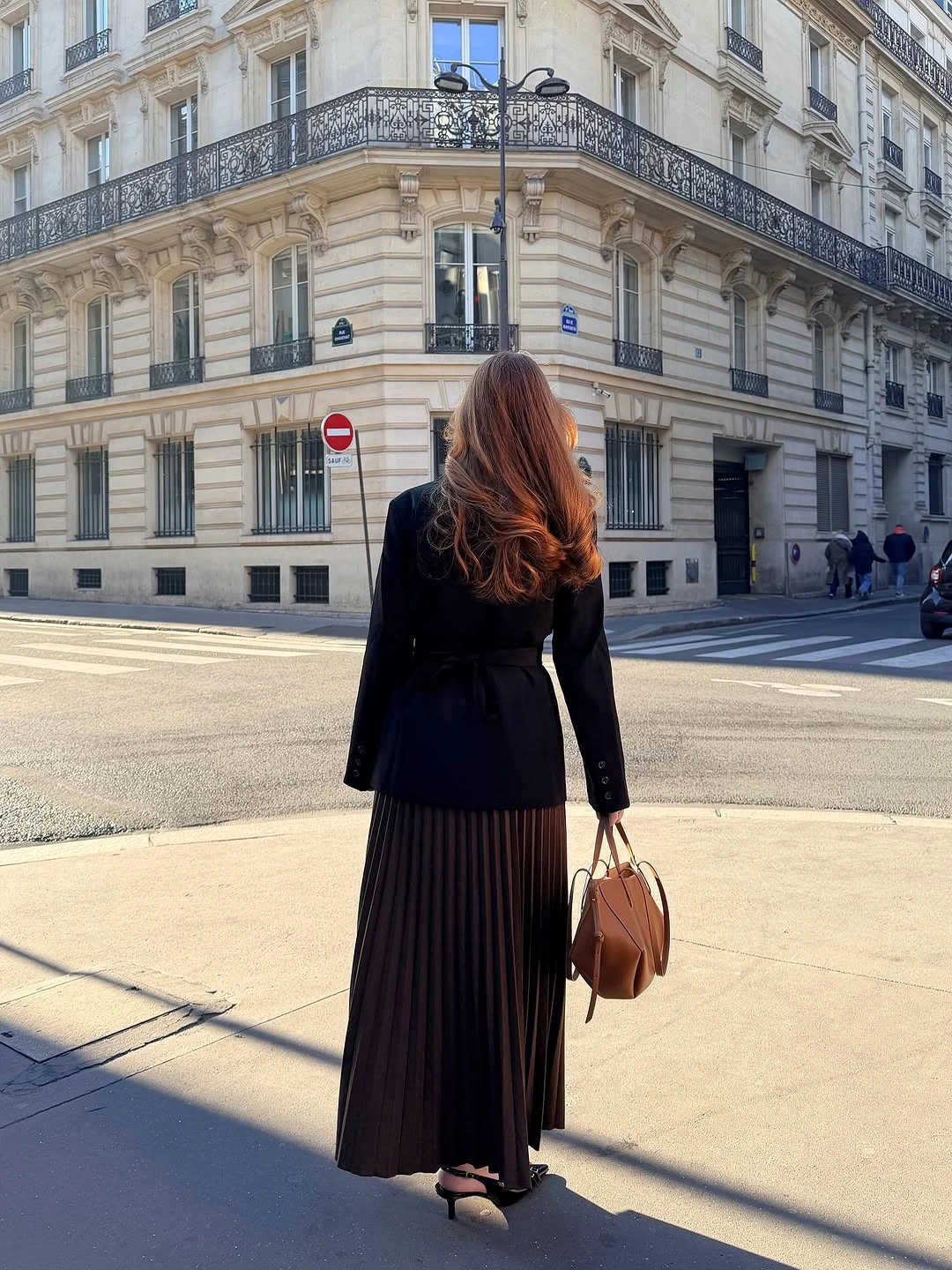 Fitted black jacket, long pleated skirt, slingbacks, and brown handbag